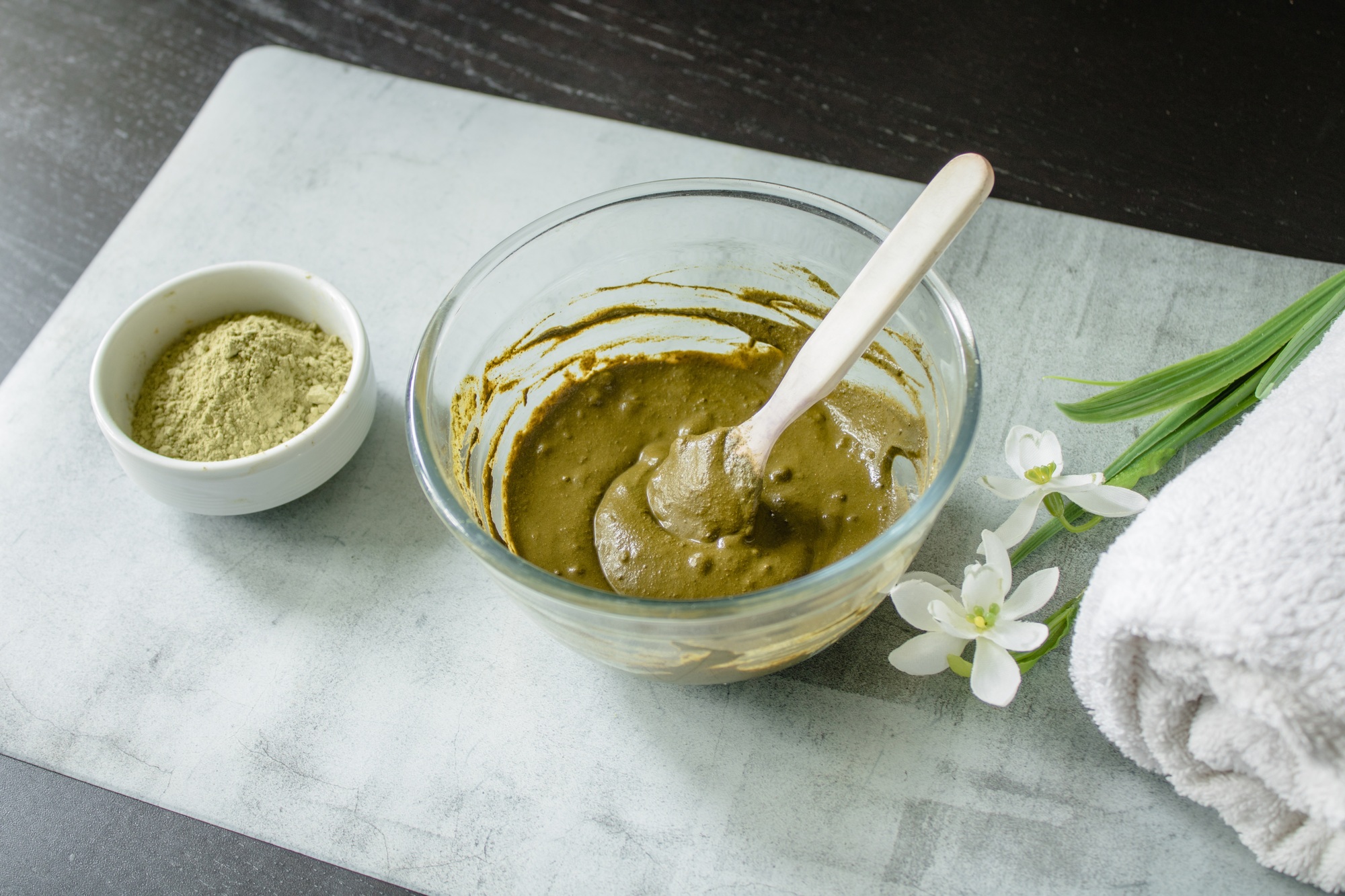 Henna dry powder, glass bowl with henna rehydrated, white terry towel on a marble mat.
